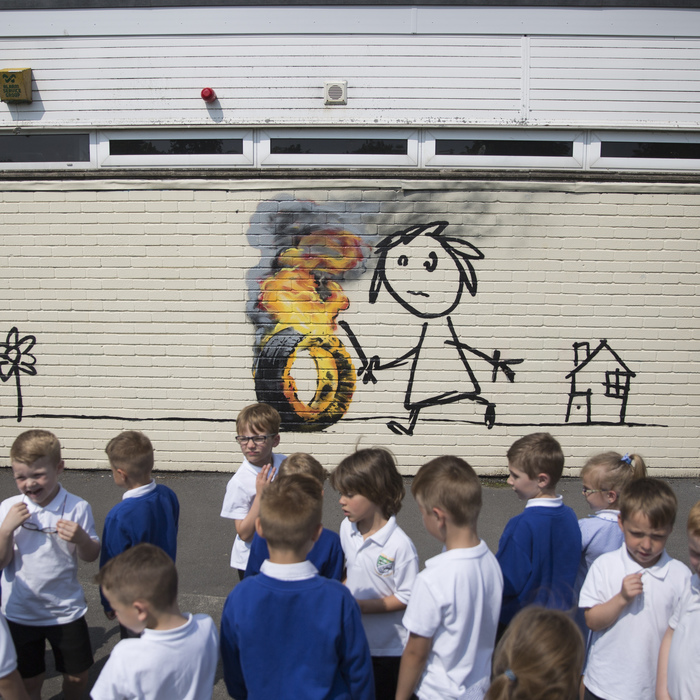 Students at the Bridge Farm Primary School in Hartcliffe, Bristol, gather round anew Banksy mural drawn recently inside the school property. June 6 2016. See SWNS story SWBANKSY: Pupils and teachers returned to their school after half-term to find a genuine BANKSY on the wall of classroom. The famous artist spray painted a stick-wielding child chasing a burning tyre on the side of Bridge Farm Primary School. It is believed the work is a modern take on hoop rolling, a popular game played by children during the Victorian days. The six-foot high artwork also features a flower and a small house with 'Banksy' signed to the bottom left of the brick building.